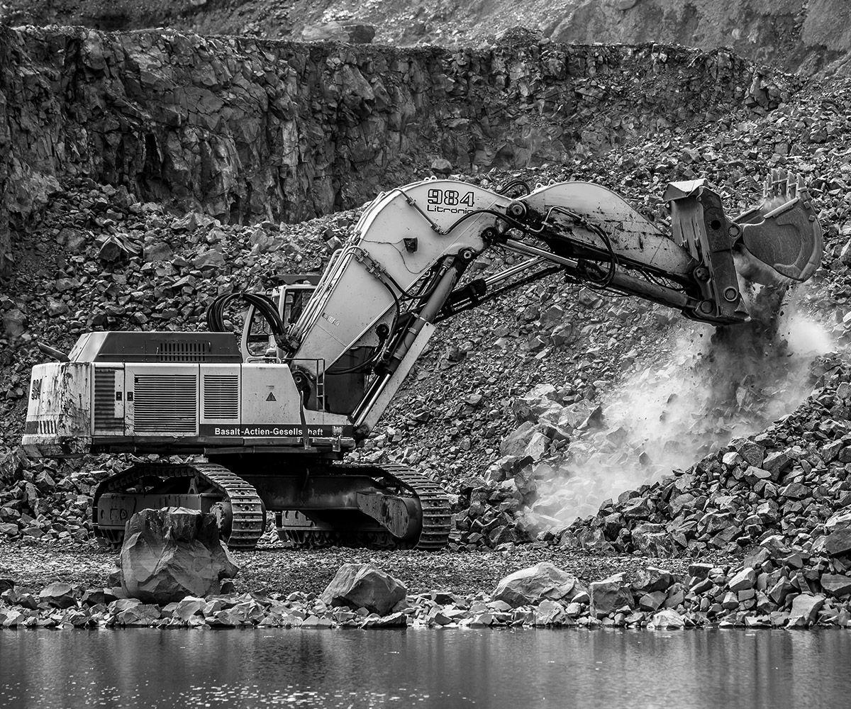 Black and white image of an excavator moving rocks by water.